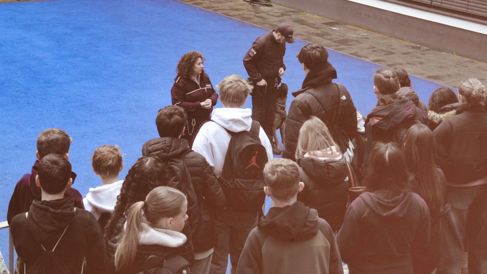 Schülerinnen und Schüler hören dem Vortrag der Diensthundestaffel zu und sehen einen trainierenden Diensthund